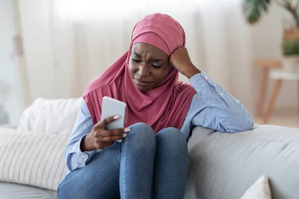 Mujer islámica negra molesta en Hijab sentada en el sofá en casa con teléfono inteligente, mirando a la pantalla del teléfono —  Fotos de Stock