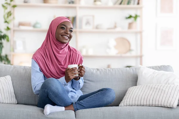Hazai pihenés. Álmodozó mosolygós fekete muszlim nő pihentető kanapén kávéval — Stock Fotó