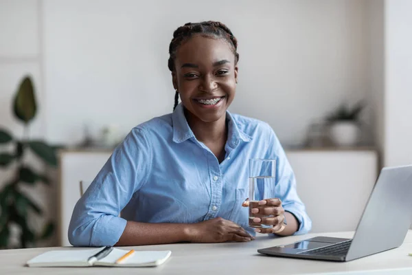 Feliz joven afroamericana sosteniendo un vaso de agua, sentada en el lugar de trabajo —  Fotos de Stock