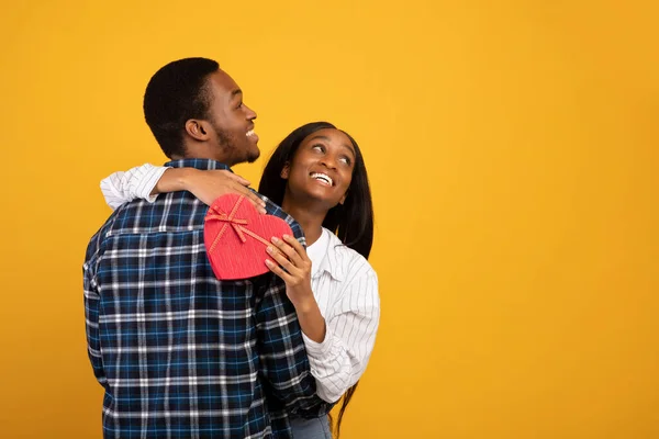 Smiling african american guy and lady hugging, look up at empty space, woman holds gift box in shape of heart