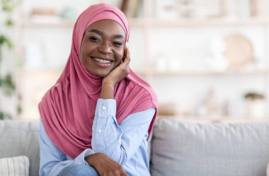 Evde kanepede oturan tesettürlü gülümseyen zenci kadın portresi.