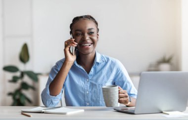 Genç siyahi kadın ofis çalışanı işyerinde cep telefonu ve kahveyle dinleniyor.