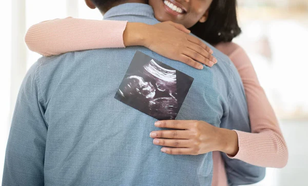 Młoda czarna kobieta w ciąży ze zdjęciem nienarodzonego dziecka przytulająca męża w domu, zbliżenie — Zdjęcie stockowe