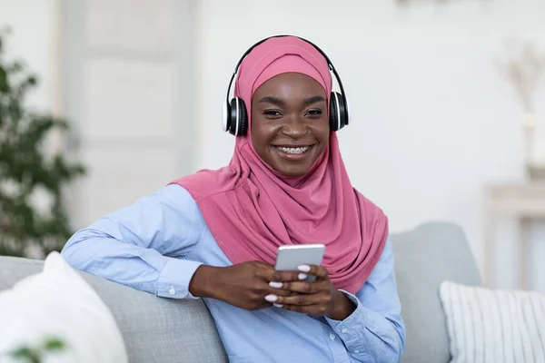 快乐的黑人穆斯林女性，带着耳机，在家用智能手机听音乐 — 图库照片