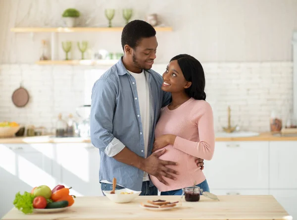 Щасливий чорний хлопець обіймає свою гарненьку вагітну дівчину і торкається її живота на кухні — стокове фото