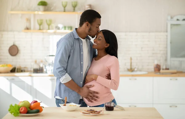 Bonito homem negro abraçando sua bela esposa grávida e beijando-a na testa na cozinha — Fotografia de Stock