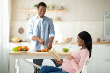 Erkek arkadaşı mutfakta kahvaltı hazırlarken siyahi hamile kadın kitap okuyor.
