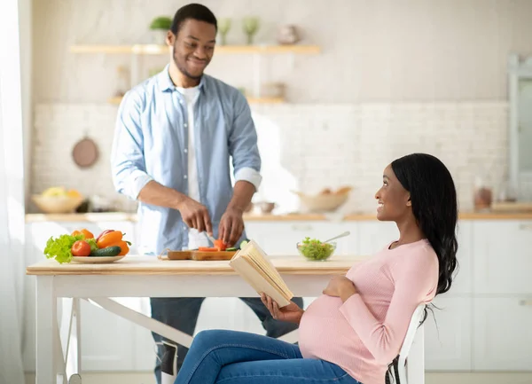 Черная беременная женщина читает книгу, пока ее муж готовит здоровый завтрак на кухне — стоковое фото