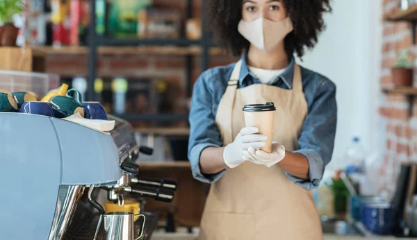 Africano americano feminino hipster garçonete em máscara facial e luvas detém takeaway bebida ordem em mãos dando xícara no café — Fotografia de Stock