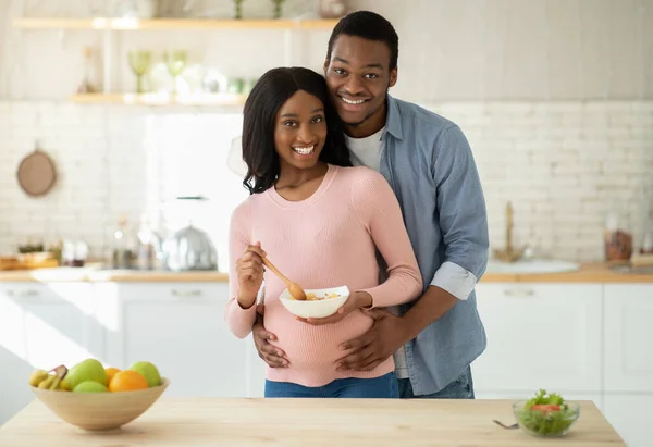 Étrend az egészséges terhességért. Pozitív fekete srác ölelgeti a kedves várandós feleségét kiegyensúlyozott étellel a konyhában. — Stock Fotó