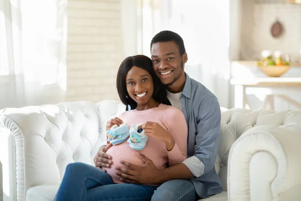 Чёрная беременная женщина с крошечными детскими ботинками и счастливым мужем, обнимающимся дома на диване. — стоковое фото