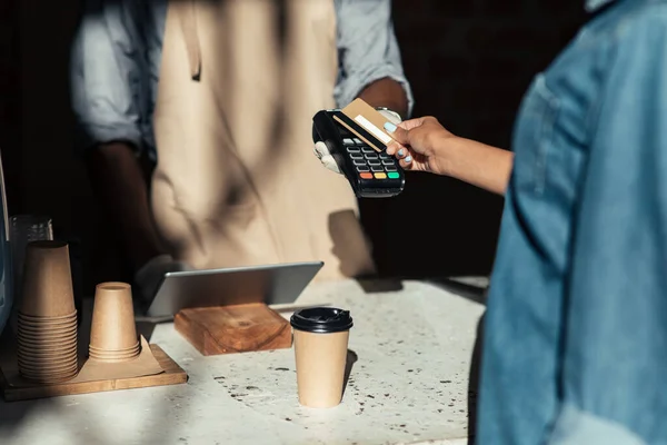 Client pays to coffee bill, millennial african american bartender in apron gives terminal in cafe counter with equipment