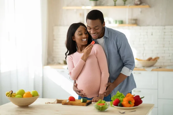 Linda mulher grávida negra alimentando seu marido com legumes enquanto cozinha na cozinha — Fotografia de Stock
