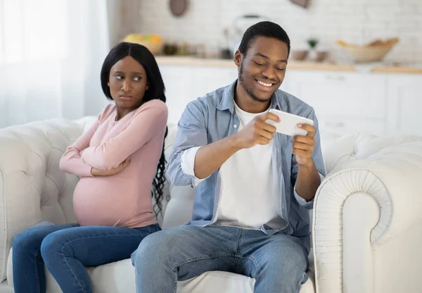 Adicción al juego móvil. Joven hombre negro jugando videojuego en el teléfono inteligente y descuidando a su esposa expectante en casa — Foto de Stock