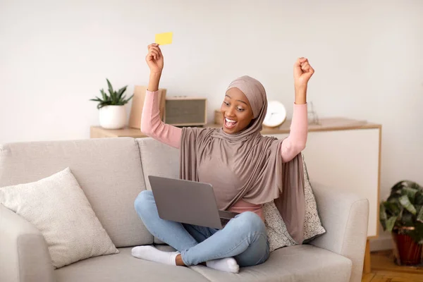 Big sale concept. Excited black woman in hijab with credit card and laptop computer shopping online at home