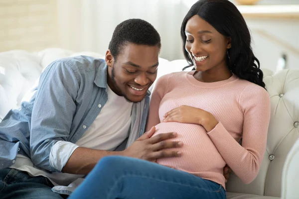 Positiver schwarzer Mann, der den Bauch seiner schwangeren Freundinnen berührt, mit dem Baby kommuniziert, seine Bewegungen zu Hause spürt — Stockfoto