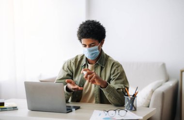 Koronavirüs sırasında uzak eğitim. Tıp maskeli siyahi genç öğrenci evde dizüstü bilgisayarın yanında antiseptik uyguluyor.