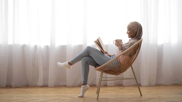 Mulher árabe preto no tradicional hijab relaxante em cadeira aconchegante com café e livro interessante em casa, espaço livre — Fotografia de Stock