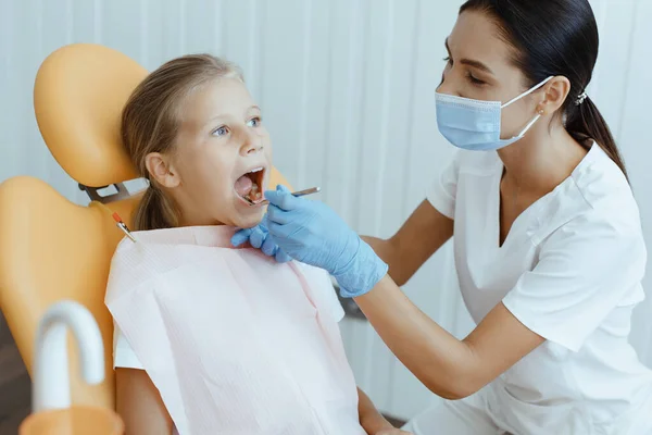 Little girl open her mouth for treatment, young female doctor in white protective mask and gloves — Stock Photo, Image