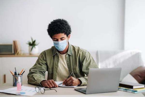 Adolescente negro em máscara médica estudando on-line no computador portátil, escrevendo notas no caderno, espaço livre — Fotografia de Stock