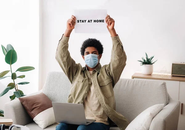 African American teenager in medical mask lifting STAY AT HOME sign above his head, indoors