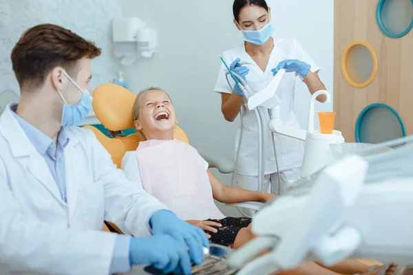 Niña riéndose en silla de dentista mira estomatología, médico joven y enfermera preparándose para el tratamiento — Foto de Stock