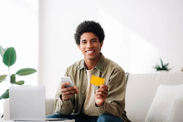 Feliz chico adolescente negro con ordenador portátil utilizando el teléfono celular y la tarjeta de crédito para las compras en línea en el interior — Foto de Stock