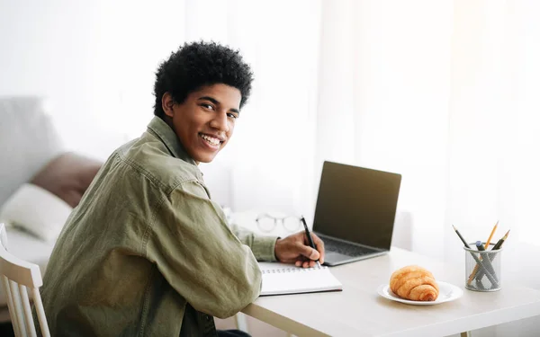 Ensino à distância. Sorrindo preto adolescente estudante escrever notas enquanto assiste vídeo educativo no laptop, espaço de cópia — Fotografia de Stock