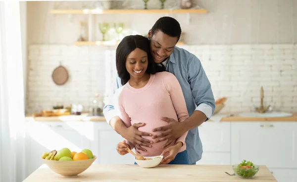 Mulher grávida muito negra cozinhar café da manhã saudável na cozinha e seu namorado abraçando-a por trás — Fotografia de Stock