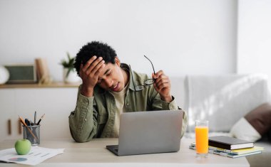 Evdeki bilgisayarın önünde online çalışmalar sırasında baş ağrısı çeken yorgun siyahi bir adam.