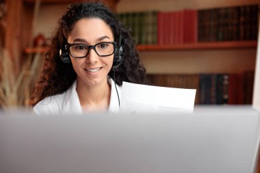 Gözlüklü kadın masada oturuyor, bilgisayarda video görüşmesi yapıyor.