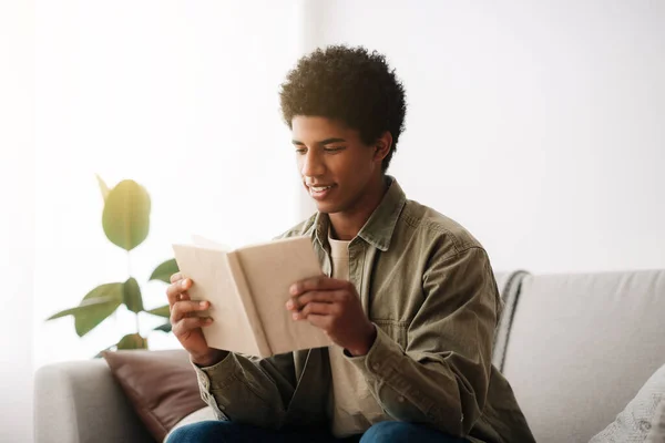 Sorridente adolescente nero che legge il libro di testo mentre è seduto sul divano a casa — Foto Stock