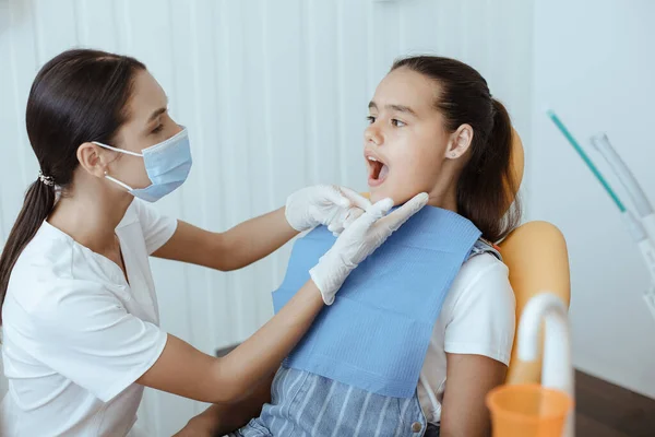 Giovane medico maschio in uniforme bianca, maschera protettiva e guanti di gomma esamina i denti della bambina — Foto Stock