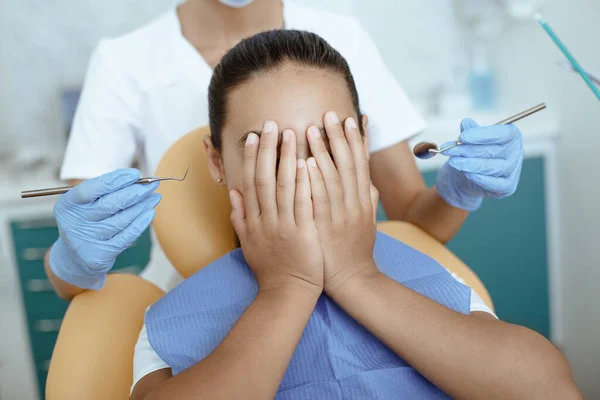 Chica asustada cubre la cara con las manos y no permite que el médico para tratar la boca con instrumentos médicos en las manos — Foto de Stock