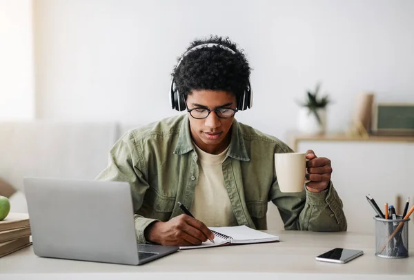 Ocupado adolescente cara bebendo café durante a aula on-line no laptop, escrevendo informações no notebook, dentro de casa — Fotografia de Stock