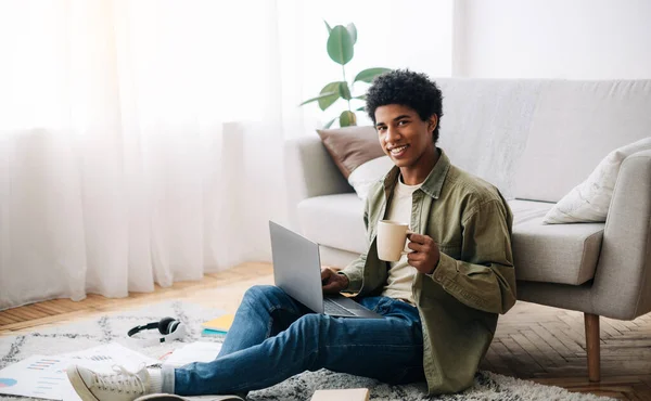 Estudante universitário preto positivo com xícara de café e laptop ouvindo palestra on-line em casa, espaço para texto — Fotografia de Stock