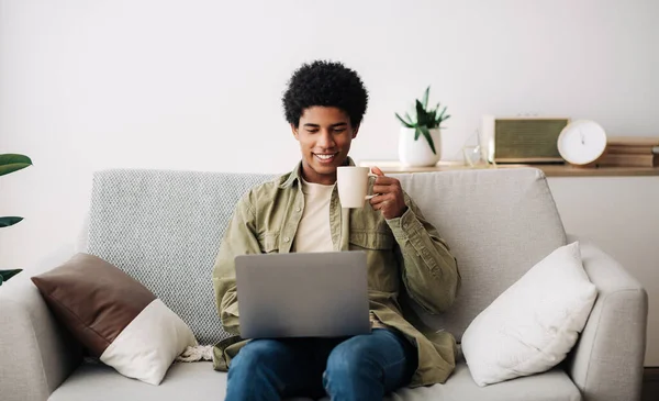 E-learning concept. Handsome black male student using laptop computer and drinking hot coffee at home, panorama — Stock Photo, Image