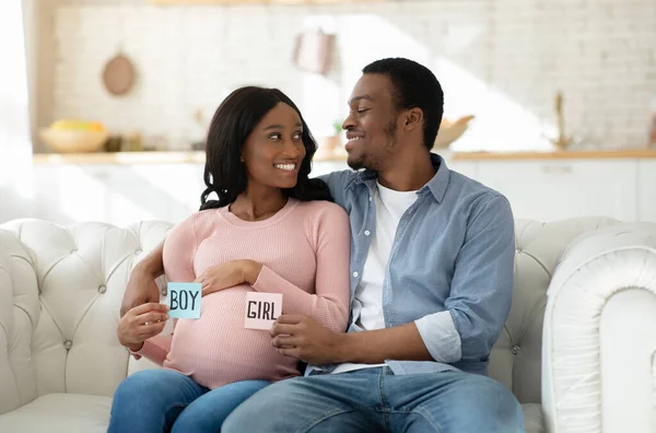 积极的黑人夫妇坐在沙发上，手里拿着纸BOY和GIRL卡，面带微笑地望着对方 — 图库照片