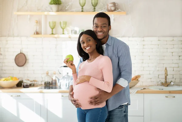 Retrato de homem negro feliz abraçando sua bela esposa grávida com fruta de maçã na cozinha — Fotografia de Stock
