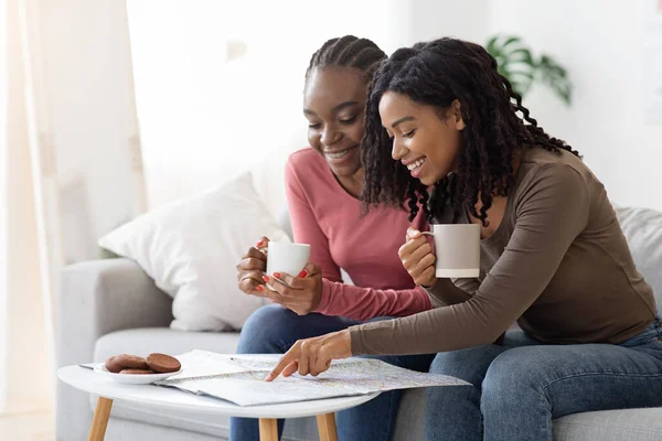 Two happy black girlfriends drinking tea and looking at map