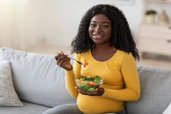 Feliz embarazada negra sentada en el sofá, comiendo ensalada fresca — Foto de Stock