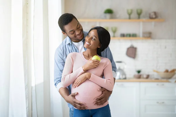 Положительная беременная черная пара обнимается, стоя рядом с окном на кухне — стоковое фото
