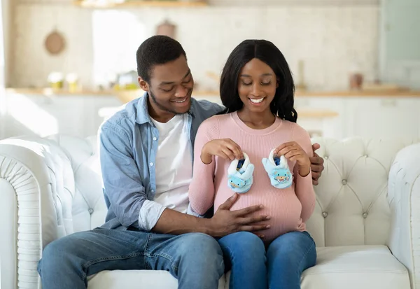 Feliz casal preto esperando novo filho, sentado no sofá juntos e segurando baby booties — Fotografia de Stock