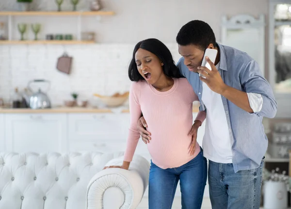 Mulher afro-americana grávida fazendo contrações pré-natais, marido preocupado chamando médico no smartphone — Fotografia de Stock