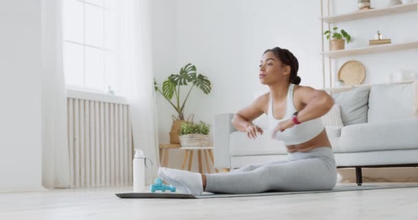 Flexibiliteitstraining. Jonge zwarte dame stretching thuis, zitten op de vloer en reiken naar de voeten — Stockvideo