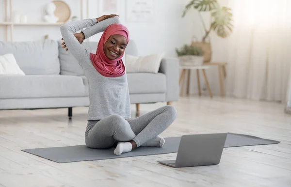 Здоровый образ жизни. Черная мусульманка делает фитнес гимнастика с ноутбуком дома — стоковое фото