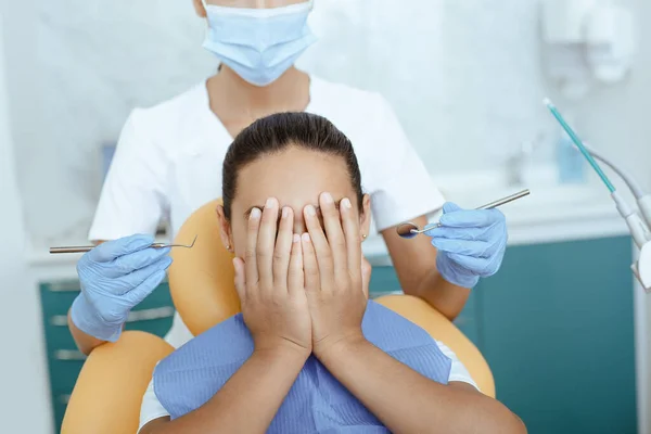 Assustado pouco latina menina no dental cadeira cobre rosto e não dá jovem mulher médico dentes exame na clínica — Fotografia de Stock