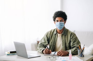 Covid-19 sırasında web tabanlı eğitim. Cerrahi maskeli siyahi bir genç dizüstü bilgisayar dersi sırasında not alıyor.