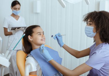 Mutlu genç doktor ve küçük kız iletişim kuruyor, Stomatolojistin portresi ve arka planda çalışan ekip