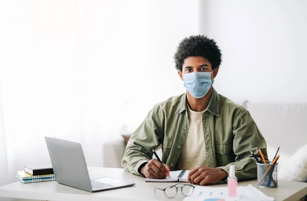 Educação baseada na Web durante o covid-19. preto teen cara no cirúrgico máscara tomando notas durante on-line palestra no laptop — Fotografia de Stock